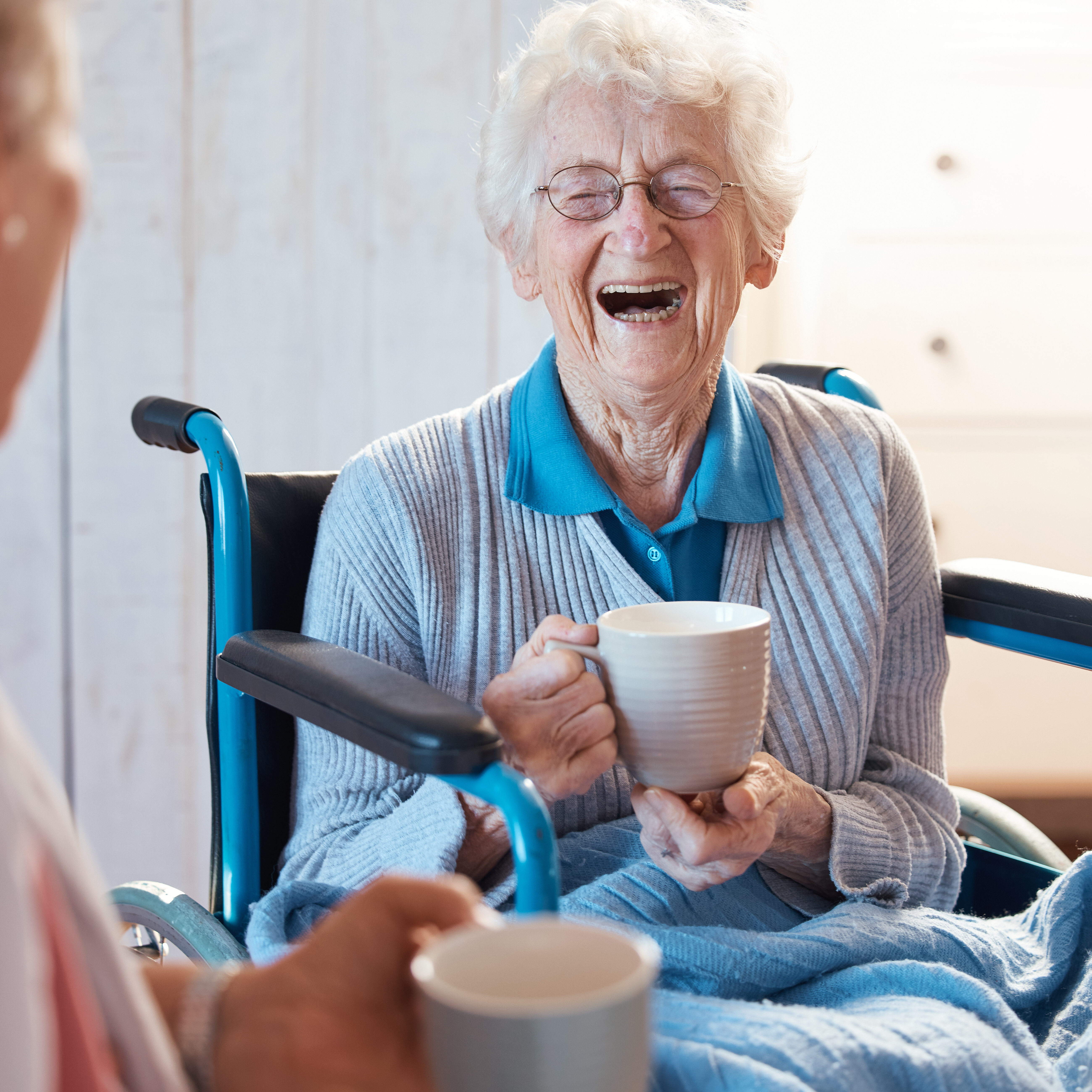 Senior woman, friend and coffee or tea while in a wheelchair for a disability or rehabilitation and happy or laughing about funny conversation or joke. Old people together for fun in nursing home.